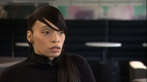 Synthia nodes in an office. She is giving an interview and looking away from the camera. The background is blurred but has seats and a table behind it. 