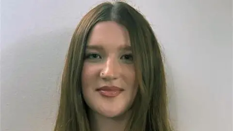 A headshot of a young woman with brown hair and a nose ring, standing with a white wall behind her.