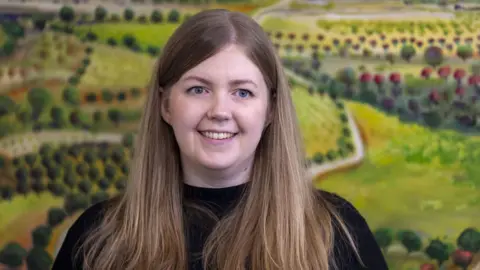 PA Media Gillian Mackay has long straight brown hair and is smiling at something out of frame 