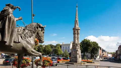 A bronze statue of a woman riding a horse at the edge of roundabout. There are planters of colourful orange, yellow and red flowers around another tall statue on the roundabout