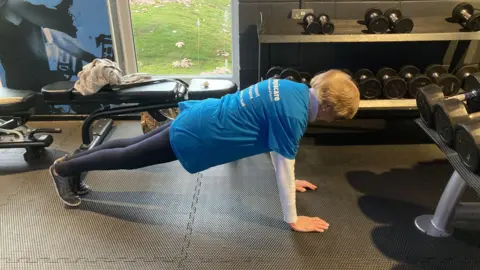 Dorrie is doing a plank with perfect form. She is wearing navy leggings and a blue T-shirt. There are racks of dumbbells of different sizes in the background.