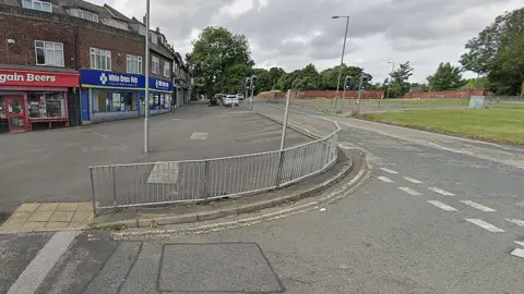 Google Muirhead Avenue in West Derby showing a row of shops and trees lining the road and traffic lights