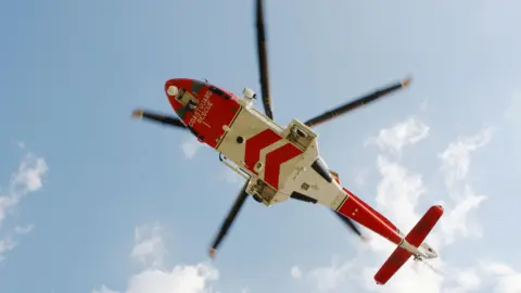 A view from the ground up towards a coastguard helicopter as it flies overhead. The aircraft has red and white markings and is seen against a blue sky with fluffy white clouds.