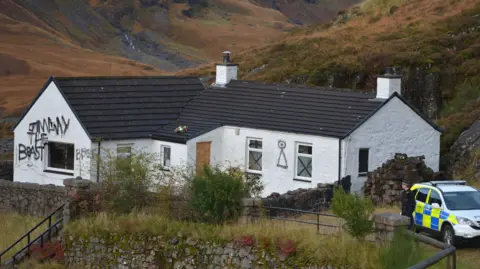 Getty Images Exterior of the main cottage at Allt Na Reigh taken in 2012 - a white single story building with a black roof with spray painted graffiti on the walls. A police vehicle and officer stands outside.  