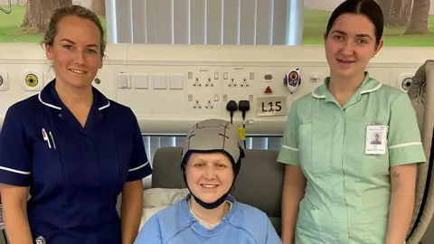 ELHT Nurse Jo Dixon is photographed in a hospital setting. She is wearing a navy blue uniform standing next to patient Lisa, who is sitting on the bed in a grey scalp-cooling cap and wearing a pale blue sweatshirt. Standing to the right in a pale green nurse's uniform is care assistant Skye Lees.