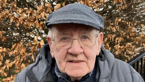 Shows an elderly man with a grey cap, grey coat and glasses