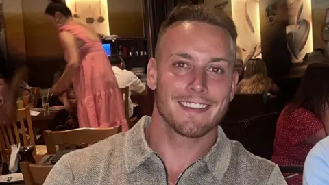 PA Media George, a man with short light brown hair and wearing a grey collared top with herringbone design fabric is smiling. He is sitting at a table in a restaurant.