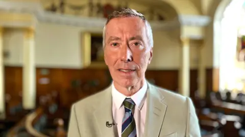 A man in a light beige jacket, white shirt and tie is looking into the camera. Behind him in a blurred background is a wood-panelled council chamber with pale yellow walls.