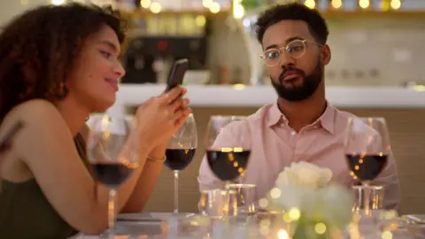 Getty Images A young couple sit at a restaurant table with glasses of wine whilst the woman is using her mobile phone and the man is looking bored and dejected.
