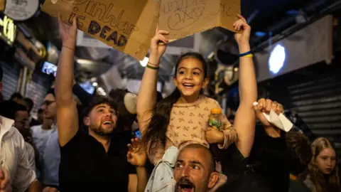 Getty Images People cheer and hold placards as they celebrate the news of the death of Hamas leader Yahya Sinwar, in Jerusalem. A man has a smiling young girl on his shoulders. People behind him wave smartphones and signs in the air.