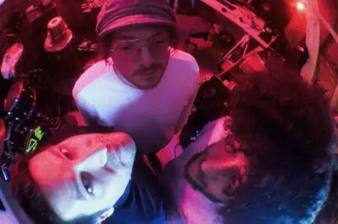 MADMADMAD Three people looking up at a fish-eye camera lens. There are musical instruments in the background. The image has a reddish tinge.