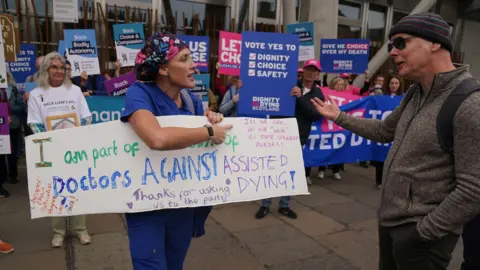 A doctor holding a sign opposing assisted dying legislation in front of supporters of the law as she argues with a man.