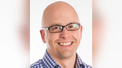 A bald man wearing glasses and a blue and white checked shirt. He is smiling towards the camera.