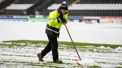 SNS A man wearing a hi-vis jacket and a bobble hat, clears snow off a football pitch with a shovel