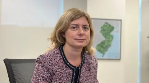 Deputy Lyndsay Feltham, a woman with shoulder-length light-brown hair, wearing a pink blazer. She is staring directly at the camera, while sitting in an office. A map is visible over her shoulder.