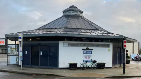 A former bus station. It is a low, possibly octagonal building with a white wall, with a bicycle resting against it, and black doors. Its black roof rises into a pinnacle. Beneath the roof and on the white wall it says "Welcome to Mildenhall High Town".