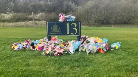 BBC/Mark Ansell A landscaped grassy area with a large dark sign standing in the center. The sign displays the number “13” in large yellow lettering and includes a smaller emblem and text reading “Owston Hall” beneath a drawing of a building. The sign is positioned upright on the grass. In front of and around the base of the sign, there is a large collection of floral tributes laid directly on the grass. The flowers are arranged in bouquets and clusters, featuring a wide range of colors including pink, yellow, white, blue, and purple. Some of the bouquets are wrapped in pastel-colored paper or clear plastic. The background consists of an open green field leading to a line of leafless or early-spring trees and shrubs, suggesting the photo was taken outdoors in a rural or park-like setting.