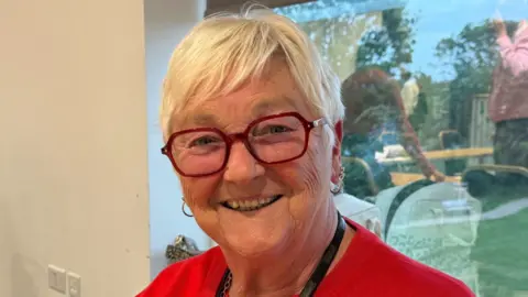 A woman - Gerri Bird - smiling straight at the camera. She is wearing a red cardigan and red glasses.
