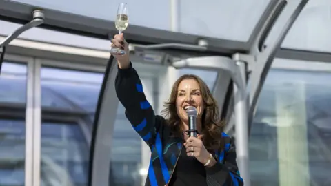 Adam Gerrard A brunette woman in a black and blue bomber jacket holds up a glass of champagne and a microphone in her other hand. She is standing in a glass pod with metallic railings.