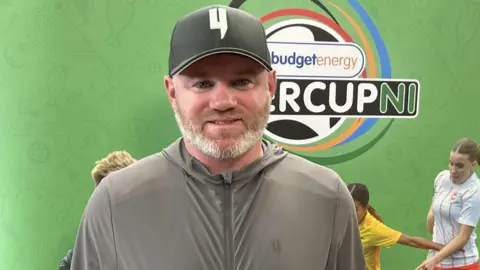 BBC Wayne Rooney wears a grey hoodie and black baseball cap. He has a white beard.
