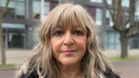 Alex Dunlop/BBC Nadia Cenci smiles at the camera. She has shoulder length blonde hair and a fringe. The background, consisting of buildings and trees, is blurred.