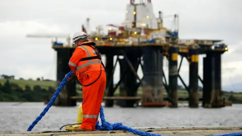 A person wearing orange reflective work gear and a white construction helmet pulls a blue rope while standing on the edge of the water as a mobile offshore drilling unit is transported into the harbour in the Port of Cromarty Firth in Cromarty in 2016.