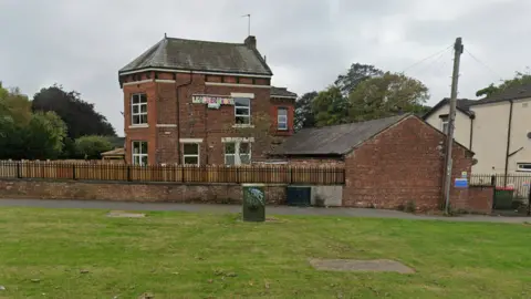 Google Leafield House Nursery on King Lane in Leeds. There is a grass verge in front of the nursery, with trees behind the red brick two-storey building. 
