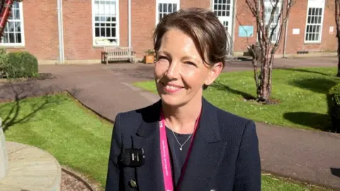 The image shows a woman with short brown hair with a pink lanyard, wearing a dark blue suit. She is standing outside a hospital building in the sunshine. 