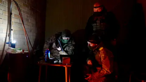 BBC/Gökte Koraltan In a dark room lit by the light of a lamp, two masked men are looking at a computer screen with another masked man standing over them.