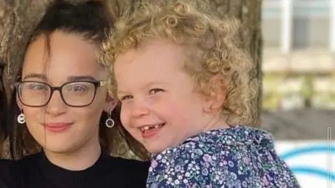 Destiny and her daughter Linnea smiling at the camera. Destiny is holding Linnea in her arms and they are smiling. 