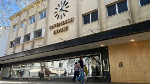 External shot of Cavendish House boarded up with a broken window. Shoppers are waking past in the foreground.