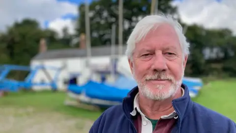 A bearded man is looking directly at the camera, dressed in a blue jacket with sailing boats behind him. 