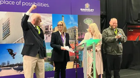 A group of four people standing on a raised stage. Behind them is a large purple backdrop featuring the text “Barnsley – the place of possibilities", along with the Barnsley Metropolitan Borough Council logo and a collage of photographs showing buildings, landmarks, green spaces, and people engaged in activities around the town.