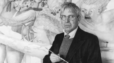 Sir Stanley, with grey hair, glasses, a paintbrush, and a dark suit, in front of his work, which includes a fierce-looking character behind him looking enraged.