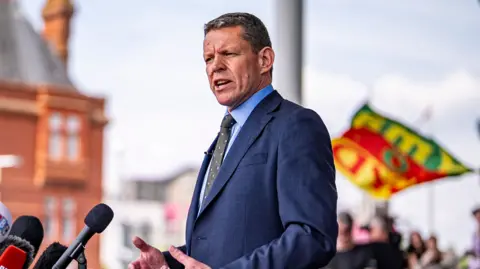 Rhun ap Iorwerth speaking into a set of microphones in front of the Pierhead building in Cardiff Bay, which is out of focus. A group of supporters can be seen behind him, also out of focus.