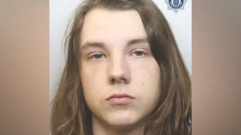 A police mugshot of a man with long brown hair. He is looking directly at the camera. He is positioned against a pale coloured background.