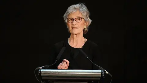 Getty Images A woman with short grey hair and glasses stands at a lectern. She is wearing a black top, and the background is black. 