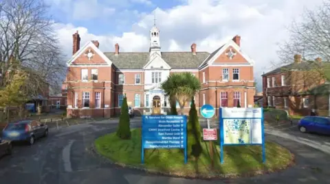 The front view of a red-brick building with a clock tower, surrounded by trees and parked cars. A blue sign in the foreground lists various services offered, and the building appears to belong to the North East London NHS Trust.