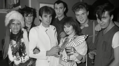 Alan Davidson/Shutterstock Black-and-white photo of a group of people posing at a party in 1981, including Spandau Ballet members Gary Kemp and John Keeble, performance duo Tik and Tok, and others.