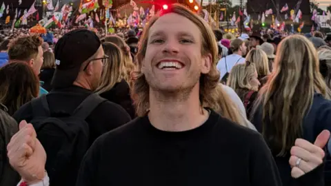 Lewis Simonds, standing in front of a crowd in Glastonbury, with his arms up. He has long fair hair and is wearing a black T-shirt. He is smiling, and has a slight beard. 