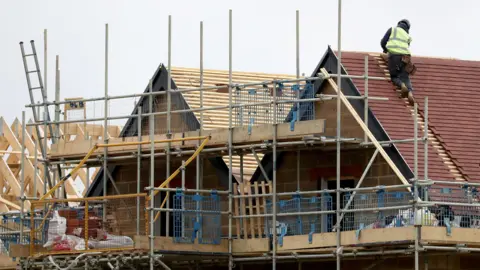 A builder works on the gable of a twin gabled house which is covered in scaffolding. They are wearing a hi-vis jacket as they climb up the slats between tiles on the roof.