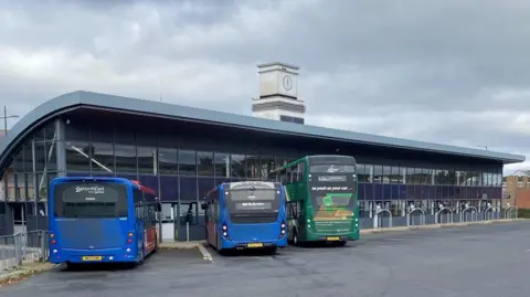 Three buses waiting at stands outside Stanley Bus Station. The long, tall building is mostly made of glass with a curved grey roof. Two of the buses are blue single-deckers. On the right is a green double-decker.