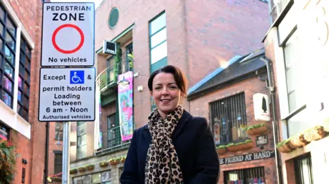 Department for Infrastructure Liz Kimmins is standing in Hill Street, Belfast. She is in front of a road sign that says pedestrian zone, no vehicles. Behind her are a collection of red brick buildings with shop signs on them. She is wearing a black coat and animal print scarf. She has medium length brown hair. 