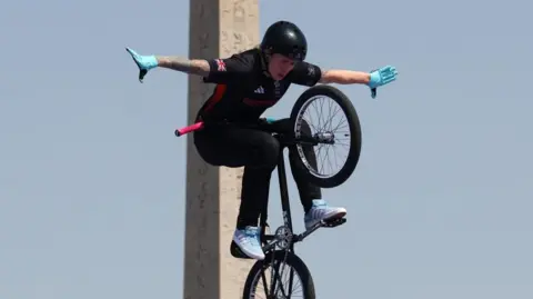 Reuters Charlotte Worthington with a black helmet and black shirt and trousers, with the Team GB logo on her sleeves. She is riding a black bike which is in the air and almost vertical alongside a stone pillar.