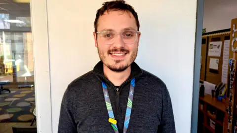 Luke smiling at the camera wearing a lanyard, dark pullover and glasses, standing in front of a wall