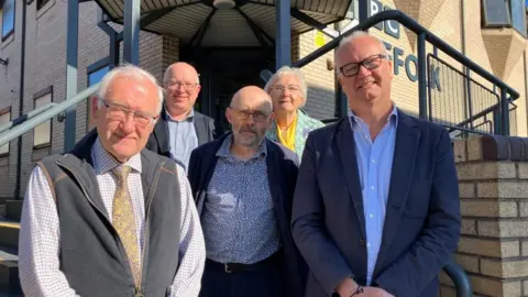The five candidates for the elections are standing on the steps in front of the BBC Radio Suffolk building. They are wearing shirts and jackets and look smart. They are all wearing glasses.