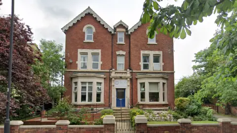 An image of the care home. It is a detached three-floor brick house with cream window frames and a front garden with grass and flowerbeds. There are trees either side of the house.