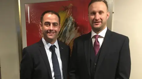 Marc Newbold Two men with short brown hair are wearing a shirt and tie and smiling at the camera. 