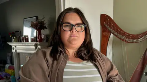 Gabriella Smith - she has long dark hair and glasses. She wears a striped top with a brown jacket over the top.