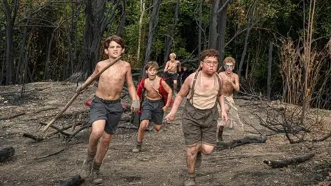 BBC/Eleven Five young boys run through a wooded area holding sticks.